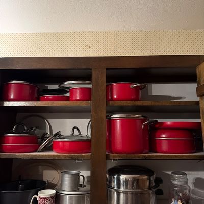 Sale Photo Thumbnail #144: Assorted vintage red enamel cookware set with lids, alongside stainless steel pots and baking accessories, displayed on wooden cabinet shelves.
