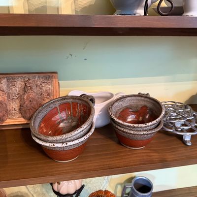Sale Photo Thumbnail #125: Collection of decorative household items arranged on a wooden shelf, featuring handmade stoneware bowls, a carved wood face plaque, a metal turtle trivet, and ceramic serving pieces.