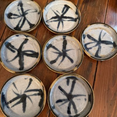 Set of seven studio pottery coasters or small plates, featuring a rustic speckled glaze with hand-painted dark blue abstract starburst...