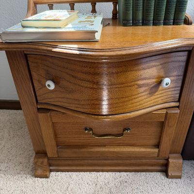 Vintage oak nightstand featuring a serpentine-front top drawer with white ceramic knobs, a lower drawer with a brass bail pull, and a...