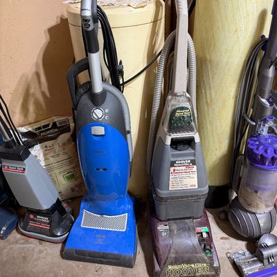 Sale Photo Thumbnail #277: A collection of floor care appliances including a bright blue Miele upright vacuum, a Hoover SteamVac Ultra carpet cleaner, a Hoover FloorMax hard floor machine, and a purple Dyson Ball upright vacuum.