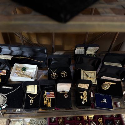 Sale Photo Thumbnail #167: Assortment of vintage and contemporary jewelry, including necklaces, rings, and patriotic flag pins, displayed in individual presentation boxes on a metal shelf.