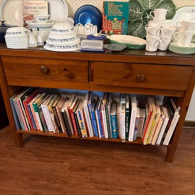 Sale Photo Thumbnail #176: Rustic pine kitchen island or console table featuring two drawers and a lower shelf filled with cookbooks, topped with an assortment of vintage dishware, mugs, and serving platters.