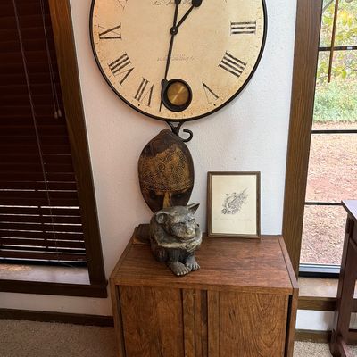 Sale Photo Thumbnail #160: Large Howard Miller wall clock with Roman numerals and a pendulum, mounted above a small wooden cabinet displaying a gargoyle statue, a decorative mask, and a framed sketch.