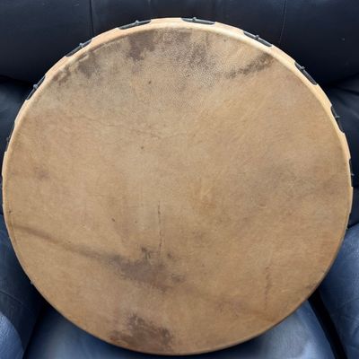 Traditional Native American style hand drum featuring a stretched rawhide head with natural coloration and lacing, resting on a blue...
