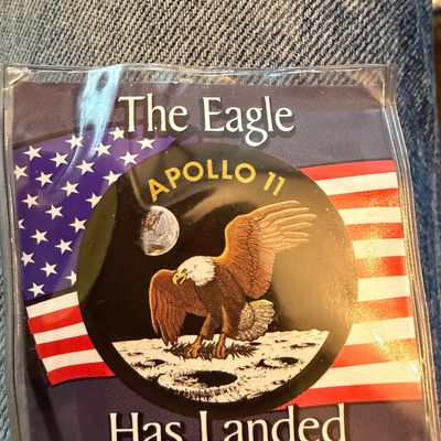 Sale Photo Thumbnail #212: Apollo 11 commemorative patch featuring the mission insignia, an American flag background, and the text "The Eagle Has Landed," preserved in a clear plastic sleeve.