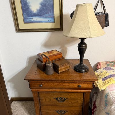Traditional oak nightstand with two drawers and brass hardware, topped with a dark-finished table lamp, decorative wooden boxes, and a...