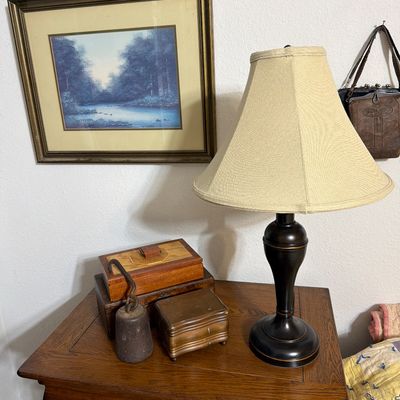 Sale Photo Thumbnail #194: Traditional dark wood table lamp with a beige fabric shade, displayed on a wooden nightstand alongside vintage wooden boxes, an antique scale weight, and a framed landscape print.