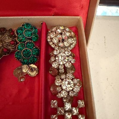 Vintage costume jewelry pieces, including a pair of green rhinestone flower earrings and a clear rhinestone brooch, displayed in a red...