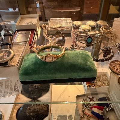 Glass display case filled with vintage jewelry and collectibles, including a gold-tone women's watch on a green velvet stand, silver...