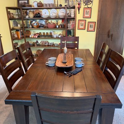 Rustic dark wood dining table with plank-style top, accompanied by six matching ladder-back chairs with dark upholstered seats.