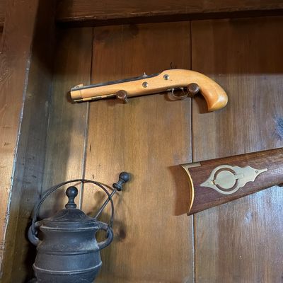 Sale Photo Thumbnail #159: Vintage replica flintlock pistol mounted on a wood panel wall, accompanied by a small cast iron kettle and the stock of a replica rifle.