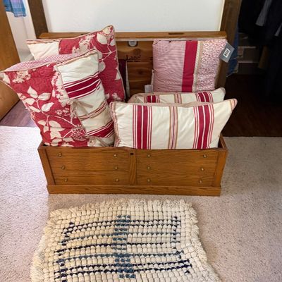 Lane cedar chest in a light oak finish, filled with assorted red and white decorative throw pillows, shown with a small textured accent rug.
