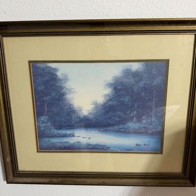 Framed landscape print depicting a serene river flowing through a dense forest, presented in a gold-toned frame with a wide beige mat.