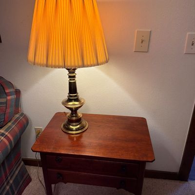 Sale Photo Thumbnail #136: Traditional brass table lamp with a pleated fabric shade, illuminated on a cherry-finished wooden side table with two drawers.