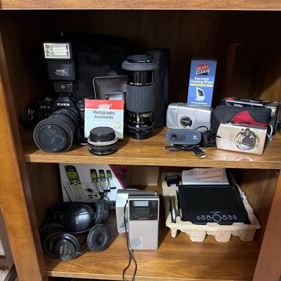 Sale Photo Thumbnail #162: Assortment of vintage and modern electronics, including a Konica FC-1 SLR camera with flash, various lenses, compact digital cameras, Sony headphones, and a portable radio displayed on wooden shelves.