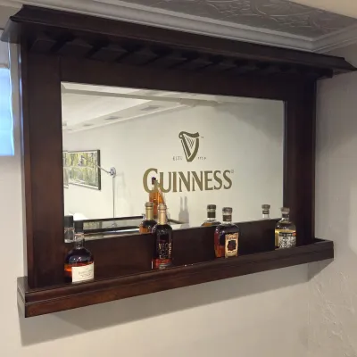 Sale Photo Thumbnail #69: Wall-mounted dark wood bar mirror featuring the Guinness logo, built-in stemware rack top, and a lower display shelf with liquor bottles.