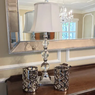 Sale Photo Thumbnail #37: Modern chrome and stacked crystal ball table lamp with a white fabric shade, flanked by a pair of silver-toned cutout decorative holders on a wooden surface.