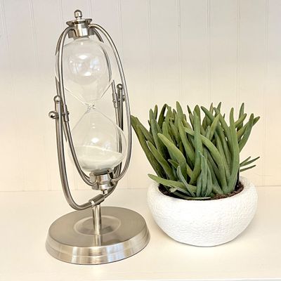 Sale Photo Thumbnail #105: Modern brushed nickel gimballed hourglass with white sand displayed next to a small potted artificial succulent.