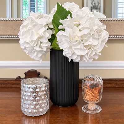 Sale Photo Thumbnail #129: Black ribbed ceramic vase filled with faux white hydrangeas, displayed alongside a mercury glass votive holder and a clear ribbed glass match cloche.