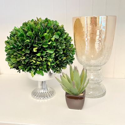 Sale Photo Thumbnail #104: Decorative tabletop trio featuring a boxwood topiary in a silver ribbed pedestal bowl, a faux potted succulent, and a tall iridescent crackle glass hurricane vase.