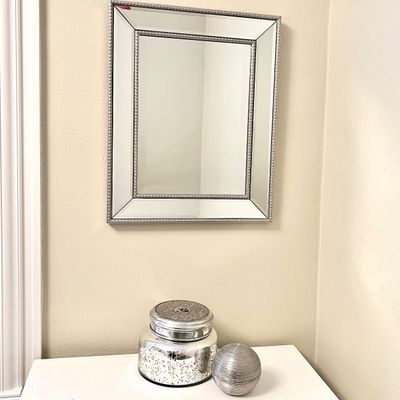 Sale Photo Thumbnail #101: Modern rectangular wall mirror with a beaded silver trim, displayed above a metallic jar candle and a ribbed silver decorative orb.