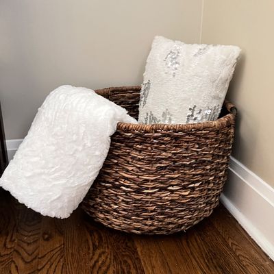 Sale Photo Thumbnail #95: Large woven dark brown wicker storage basket holding a plush white faux fur blanket and a sequined white throw pillow.