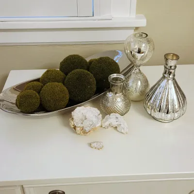 Sale Photo Thumbnail #77: Collection of decorative modern tabletop accents, including a metallic tray filled with green moss balls, mercury glass vases, a glass hourglass, and quartz geode specimens.