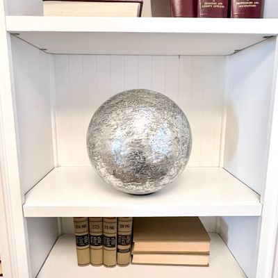 Sale Photo Thumbnail #103: Large decorative silver mercury glass orb displayed on a white bookshelf alongside vintage books.