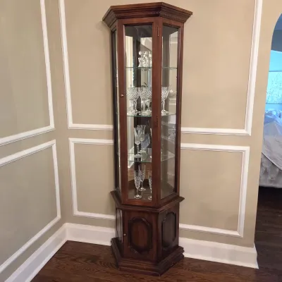 Sale Photo Thumbnail #41: Tall hexagonal wood curio cabinet featuring glass display shelves above a lower storage base, filled with cut crystal stemware.