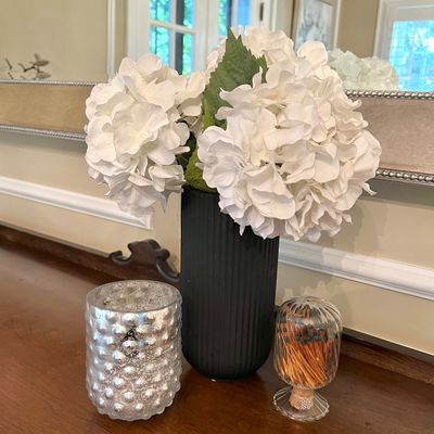 Sale Photo Thumbnail #14: A decorative tabletop arrangement featuring faux white hydrangeas in a matte black ribbed vase, flanked by a mercury glass candle holder and a glass match striker.