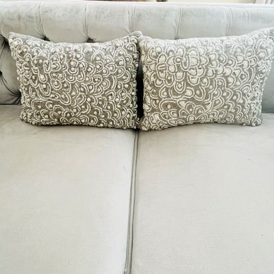 Sale Photo Thumbnail #20: Pair of contemporary beaded throw pillows with intricate white and metallic scrollwork on a neutral couch.