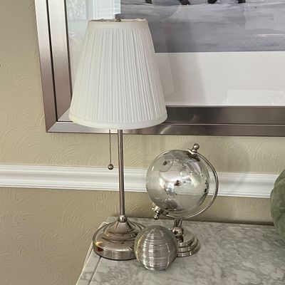 Sale Photo Thumbnail #13: Brushed nickel table lamp with a pleated white fabric shade, alongside a decorative mirrored glass globe on a metallic stand.