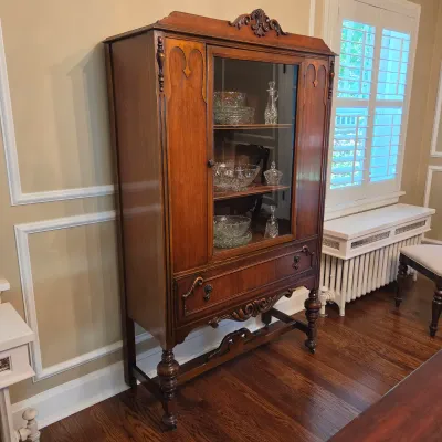 Sale Photo Thumbnail #40: Vintage Jacobean Revival dark wood china cabinet with carved pediment, glass display door, and lower drawer, displaying a collection of cut crystal glassware.