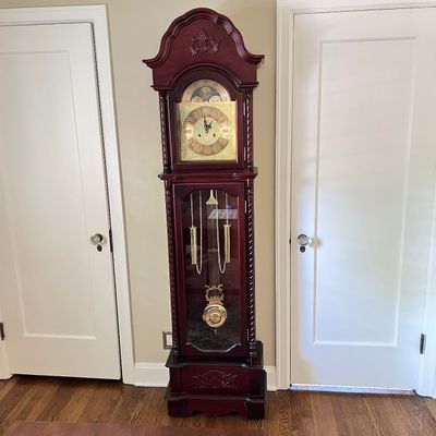 Sale Photo Thumbnail #99: Traditional cherry-finished grandfather clock featuring a bonnet top with carved detail, a brass moon phase dial, and visible weights and lyre pendulum behind a glass door.