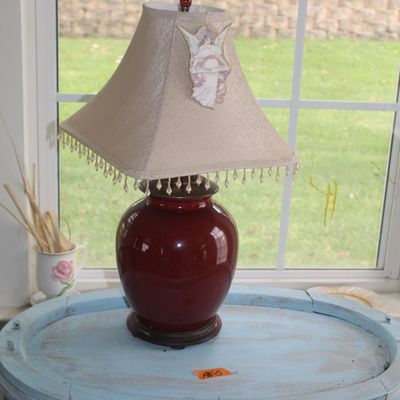 Sale Photo Thumbnail #71: Oxblood red ceramic ginger jar table lamp, featuring a beige fabric shade with beaded fringe trim and an angel ornament.