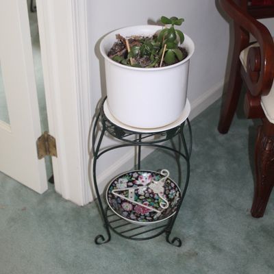 Sale Photo Thumbnail #79: Two-tier black wrought iron plant stand holding a white ceramic potted jade plant on top and a floral decorative plate on the bottom shelf.