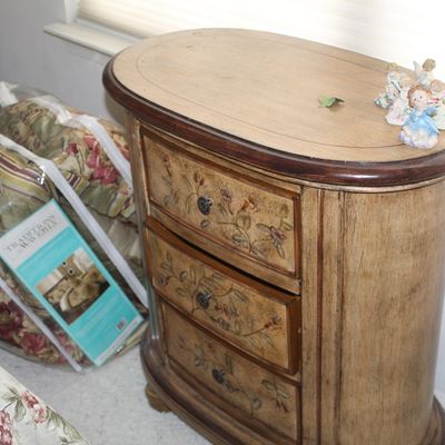 Sale Photo Thumbnail #50: Traditional oval wooden nightstand featuring three drawers with hand-painted floral details and dark metal pulls, shown alongside packaged Waverly bedding.