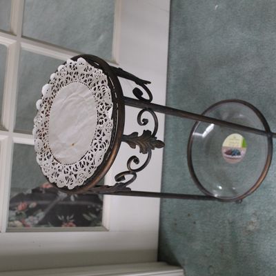 Sale Photo Thumbnail #68: Two-tier metal plant stand with scrollwork details, featuring a glass bottom shelf and a white paper doily on the top shelf.