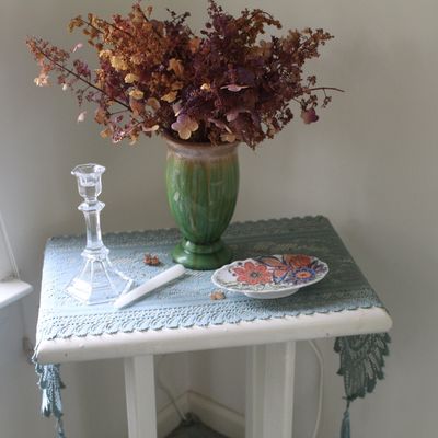 Sale Photo Thumbnail #76: Small white painted side table featuring a green glazed ceramic vase with dried flowers, a clear glass candlestick, and a small floral dish on a blue lace runner.
