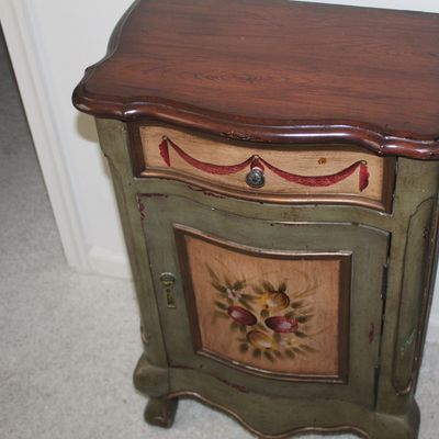 Sale Photo Thumbnail #43: Vintage hand-painted wooden accent cabinet featuring a dark wood top, a single drawer with red swag detail, and a lower cabinet door adorned with a floral motif.
