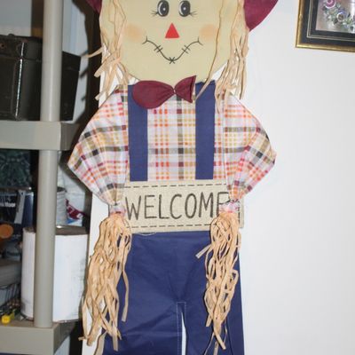 Sale Photo Thumbnail #53: Whimsical scarecrow welcome sign hanging decoration, featuring a plaid shirt, blue overalls, raffia hair, and a maroon hat with a leaf accent.