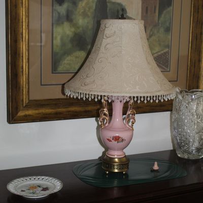Sale Photo Thumbnail #65: Vintage pink porcelain table lamp with gold-painted handles, floral motif, and a fringed fabric shade, displayed alongside a framed landscape print and a small decorative plate.