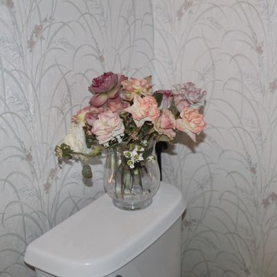 Sale Photo Thumbnail #118: Faux floral arrangement of pink and white roses in a clear glass vase, displayed on a toilet tank against patterned wallpaper.
