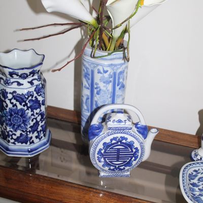 Sale Photo Thumbnail #12: Collection of blue and white Chinese porcelain items, including a cylindrical vase with faux calla lilies, a hexagonal vase, and a decorative teapot, displayed on a glass-top wooden table.