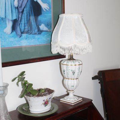 Sale Photo Thumbnail #82: Vintage porcelain table lamp with gold trim and green floral accents, paired with a fringed fabric shade, displayed alongside a potted jade plant on a wooden desk.