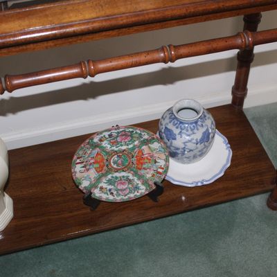 Sale Photo Thumbnail #14: Assorted Asian porcelain collectibles, including a Rose Medallion plate on a stand and a blue and white ginger jar, displayed on the lower shelf of a faux-bamboo wooden console table.