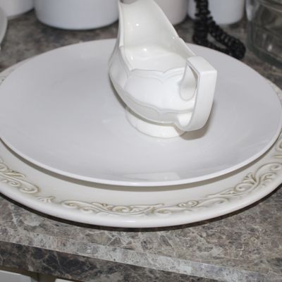 Sale Photo Thumbnail #101: White ceramic gravy boat resting on a matching white serving platter, displayed alongside a larger decorative charger plate with a raised scrollwork border.