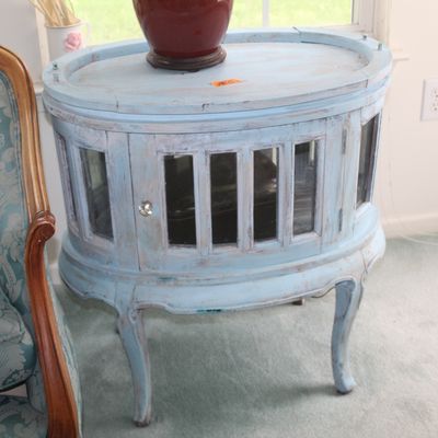 Sale Photo Thumbnail #70: Vintage oval wooden side table with a distressed light blue painted finish, featuring glass-paneled cabinet doors and cabriole legs.