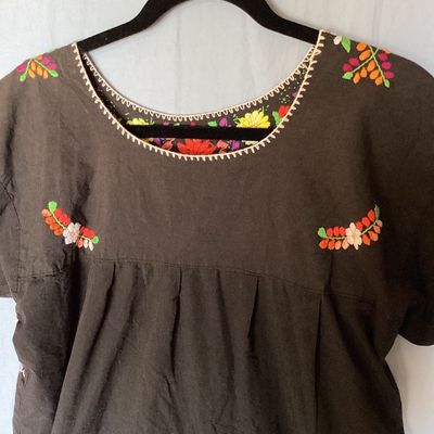 Sale Photo Thumbnail #396: Black short-sleeve embroidered Mexican huipil tunic dress featuring colorful floral embroidery across the neckline, chest, and front lower panel. Features a scoop neckline with stitch details and side vents at the hem. Measures approximately 35 inches in 